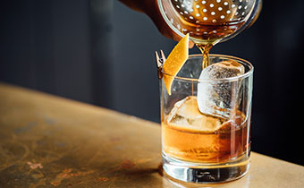 Bartender straining a cocktail into a glass with an orange peel garnish